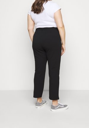 Mujer con cabello largo y castaño, que lleva una camiseta blanca, pantalones negros de tiro alto y zapatillas grises, de pie contra un fondo blanco liso.