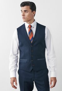Navyblauer strukturierter Weste aus Stoff, ausgestattet mit fünf Knöpfen, zwei Taschen und einem weißen Hemd mit einer gestreiften orange- und blau-roten Krawatte.