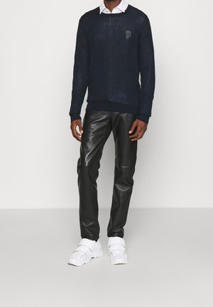 Modèle masculin portant un pull bleu marine foncé sur une chemise blanche à col, un pantalon en cuir noir et des baskets blanches, debout devant un fond uni.