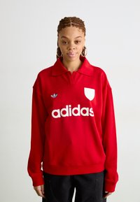 Felpa rossa in tessuto misto cotone. Presenta un grande logo bianco "adidas" e un emblema trefoil blu sul lato sinistro del petto.