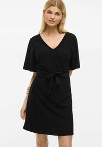Svart klänning med korta ärmar, V-ringning och ett knutet bälte i midjan. Har en texturerad tyg med subtila utskurna mönster.
