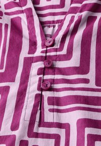 Camicia lavanda con audaci motivi geometrici bianchi, dotata di scollatura a V e tre bottoni viola sul davanti. Texture in cotone morbido.