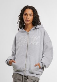 Grauer Reißverschluss-Hoodie mit großer Kapuze, Fronttaschen und gesticktem Text "Feeling Delicious" in Weiß. Weicher Stoff und lässige Passform.