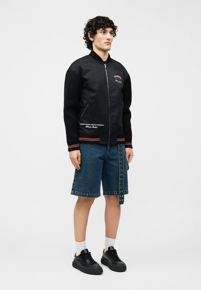 Νεαρός άνδρας με σγουρά μαλλιά, φοράει μαύρο μπουφάν με φερμουάρ και κείμενο, τζιν σορτς μέχρι τα γόνατα με ζώνη, λευκές κάλτσες και μαύρα αθλητικά παπούτσια.