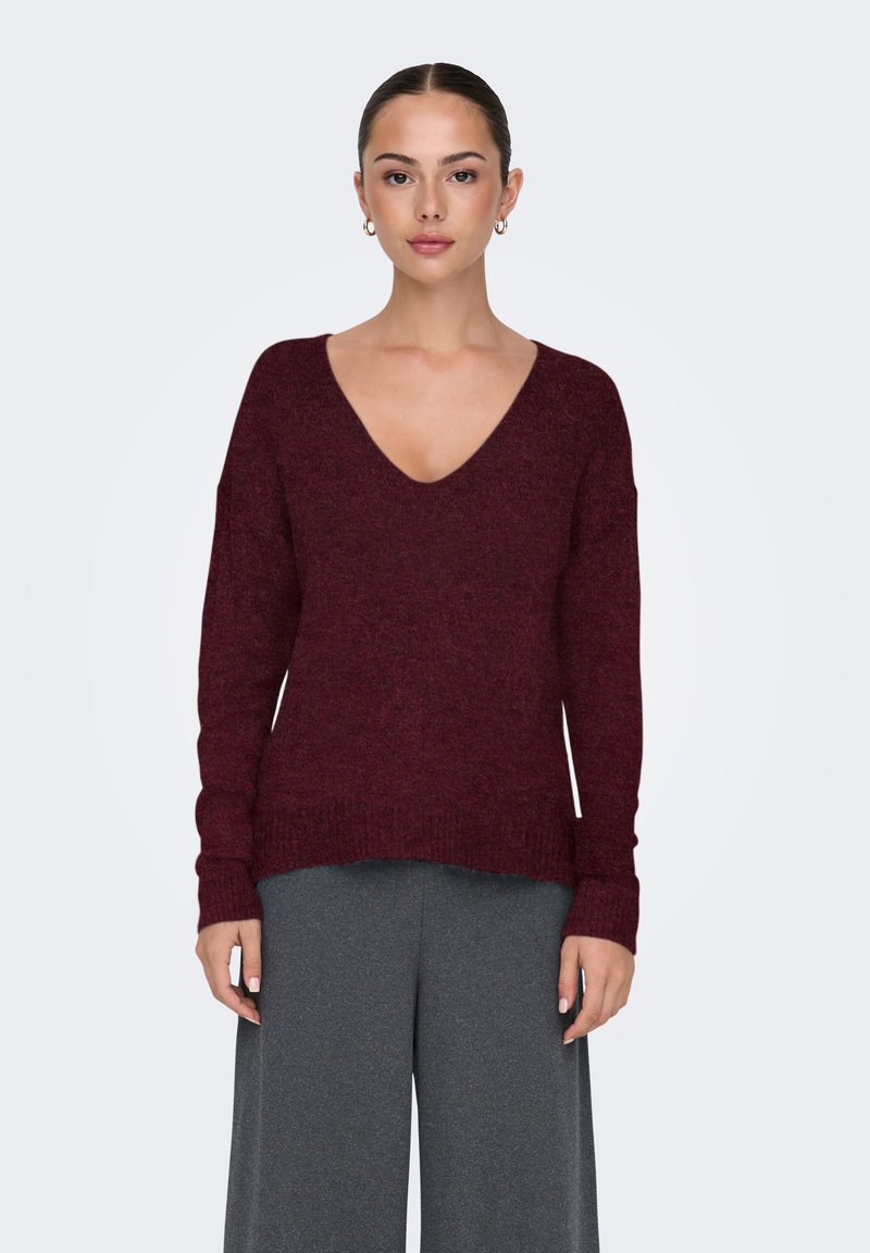 Vrouw met naar achteren gestyled haar, die een bordeauxrode V-hals trui en losse grijze broek draagt, staand tegen een eenvoudige lichte achtergrond.