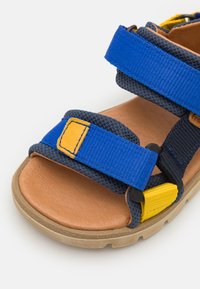Blå og gul barne sandal med justerbare stoffremmer, teksturert overflate og en brun såle; har en polstret fotbed for komfort.