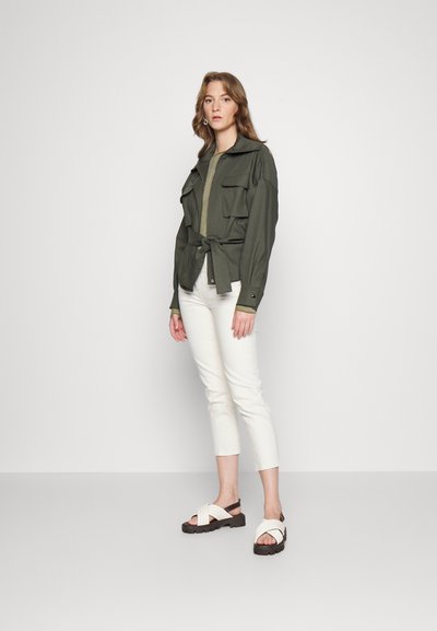 Casaco verde-oliva com cinto e dois bolsos frontais, combinado com uma blusa verde canelada e calças brancas cropped. Usado com sandálias creme de tiras.