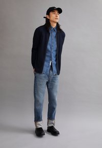 Marineblauwe rits cardigan over een denim shirt, gecombineerd met lichtblauwe jeans met opgerolde zoom. Zwarte schoenen en een zwarte pet maken de look compleet.
