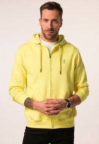 Geel rits-hoodie met trekkoorden, voorzakken en geribbelde manchetten. Bevat een klein logo op de borst. De stof lijkt zacht en dik.