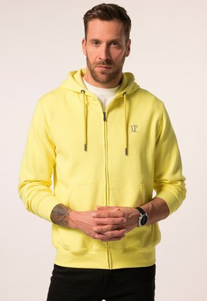 Haut zippé jaune avec cordons de serrage, poches avant et poignets côtelés. Présente un petit logo sur la poitrine. Le tissu semble doux et épais.