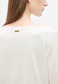 Fehér, könnyű felső kerek nyakkivágással és egy kis arany márkajelzéssel a hátoldalon, sima textúrával és laza szabással.