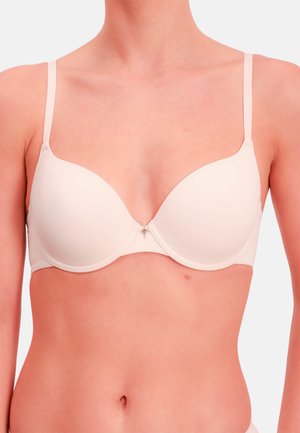 Kobieta ubrana w jasno beżowy, lekko wyściełany biustonosz z regulowanymi ramiączkami i małym ozdobnym zawieszkiem na środku z przodu.