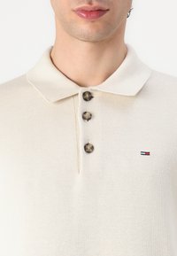 Beežist kraega polo särke, millel on kolm tumedat nuppu, ribatatud tekstuur ja väike logo punaste, valgete ja siniste aktsentidega vasakul küljel.