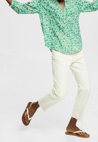 Camicia verde con stampa floreale e scollo a V; pantaloni color crema, leggermente usurati; sandali piatti marroni con cinturini bianchi.