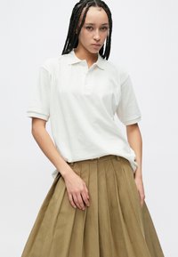 Vit pikétröja med klassisk krage, korta ärmar och en diskret varumärkeslogotyp. Matchas med en plisserad, högmidjad brun kjol.