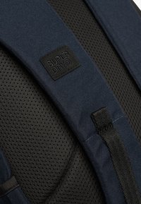 Correa de mochila de la marina con malla negra texturizada y transpirable, y un parche de cuero negro con la etiqueta "SUPERDRY". Cuenta con hebillas ajustables.