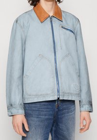 Ljusblå denimjacka med brunt läderkrage, frontdragkedja, två sidofickor och en liten bröstficka med blå dragkedja.