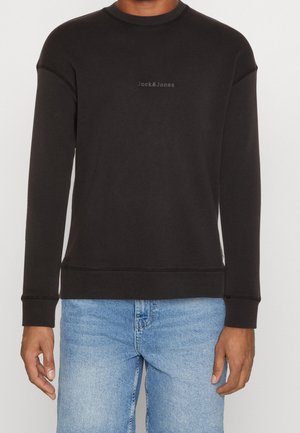 Czarna bluza z długim rękawem i okrągłym dekoltem z logo "Jack & Jones" na piersi, noszona przez osobę w jasnoniebieskich dżinsach, stojącą na neutralnym tle.