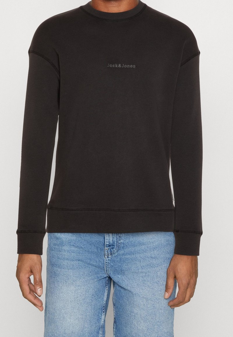 Sweat-shirt noir à manches longues col rond avec logo « Jack & Jones » sur la poitrine, porté par une personne en jean bleu clair, debout devant un fond neutre.