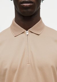 Κοντινό πλάνο στο λαιμό και το άνω μέρος του στήθους που φοράει μπεζ πόλο με υφασμένο γιακά και μερικώς ανοιχτό ασημί φερμουάρ.