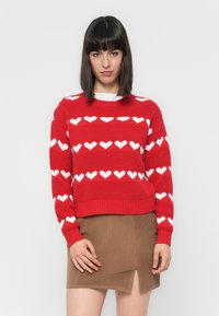 Czerwony sweter dzianinowy z białymi wzorami serc. Sweter ma prążkowane mankiety i dół. Połączony z brązową mini spódnicą z sztruksu z rozcięciem.