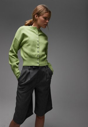 Femme aux cheveux châtain clair attachés en queue de cheval, portant un cardigan vert clair et un short gris foncé taille haute jusqu’aux genoux, les mains dans les poches.