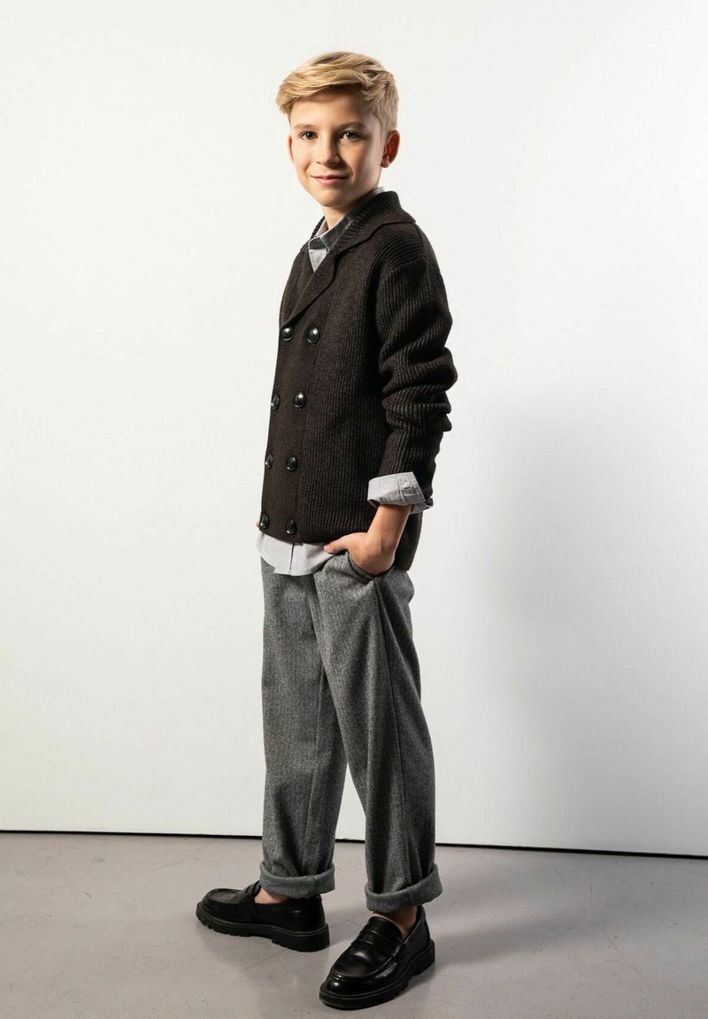 Brązowy dziergany sweter zapinany na dwa rzędy guzików, szare wełniane spodnie, czarne mokasyny, biała koszula z kraciastym kołnierzem, stojąca poza na neutralnym tle.