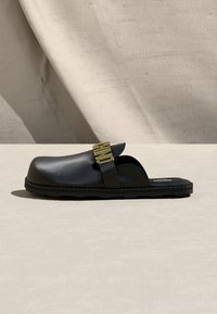 Μαύρο πάνινο παπούτσι με χρυσό λουράκι με αγκράφα και επίπεδη, ραβδωτή σόλα σε μπεζ φόντο.