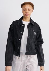 Giacca cropped in denim nera con bottoni argentati e tasca sul petto. Presenta sottili righe verticali e un colletto classico. Indossata con pantaloni a righe.