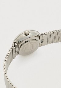 Achteraanzicht van een zilveren roestvrijstalen Tommy Hilfiger horloge met een mesh band en gegraveerde merkdetails op de achterkant van de behuizing.