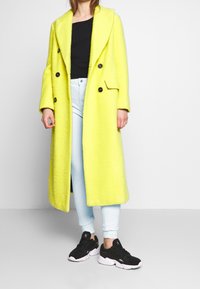 Henkilö, joka wear pitkän kirkkaan keltaisen takin, mustan paidan, vaaleansiniset farkut ja mustat urheilukengät, seisoo tasaista taustaa vasten.