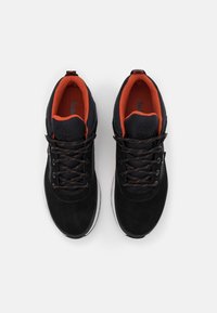 Paire de baskets noires avec des lacets noirs, des semelles blanches et une doublure intérieure orange vif, vues de dessus sur fond blanc.
