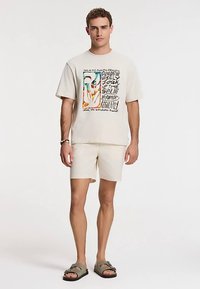 Beež värvilise abstraktse trükiga graafiline t-särk, mida on kombineeritud helbeežide lühikestega. Vabaaja riietuse lõpetavad sandaalid.