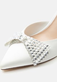 Λευκό παπούτσι με μυτερή μύτη και μεγάλο φιόγκο διακοσμημένο με διάφανα, στρογγυλά στρας στο μπροστινό πάνω μέρος.