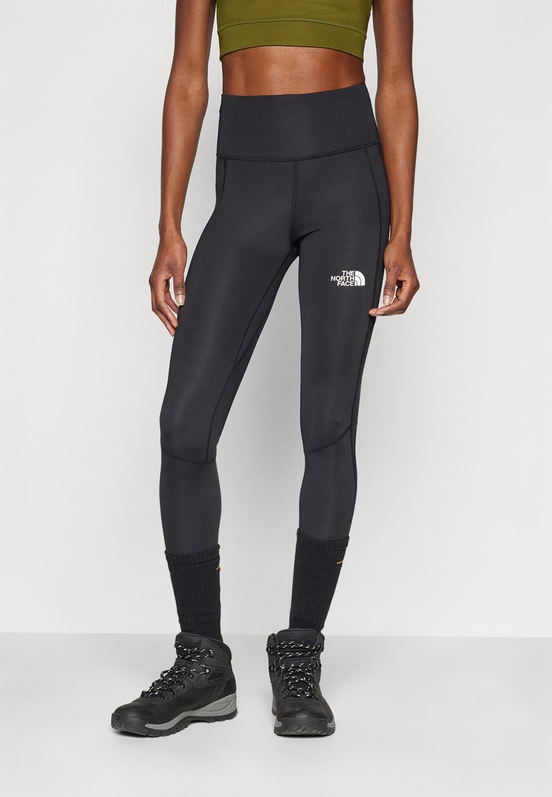 Leggings neri con vita alta, realizzati in tessuto elastico, con un logo bianco sul lato sinistro e indossati con stivali da trekking neri.