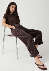 Woman with long dark hair wearing a brown t-shirt, wide brown pants, and sandals, sitting relaxed on a metal chair against a plain background.