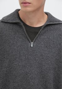 Pull gris en maille avec un col à fermeture éclair, présentant un motif texturé. Porté sur une chemise foncée, il offre un look épuré et moderne.