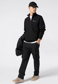 Sudadera negra con cremallera hasta un cuarto, con el logo de Champion, combinada con pantalones cargo negros. Zapatillas de color neutro. Modelo con gorra negra.