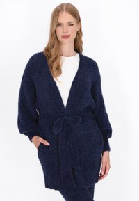 Granatowy sweter z dzianiny, z przewiązaniem w pasie, z dużymi rękawami i kieszeniami. Miękka, teksturowana tkanina o luźnym kroju.