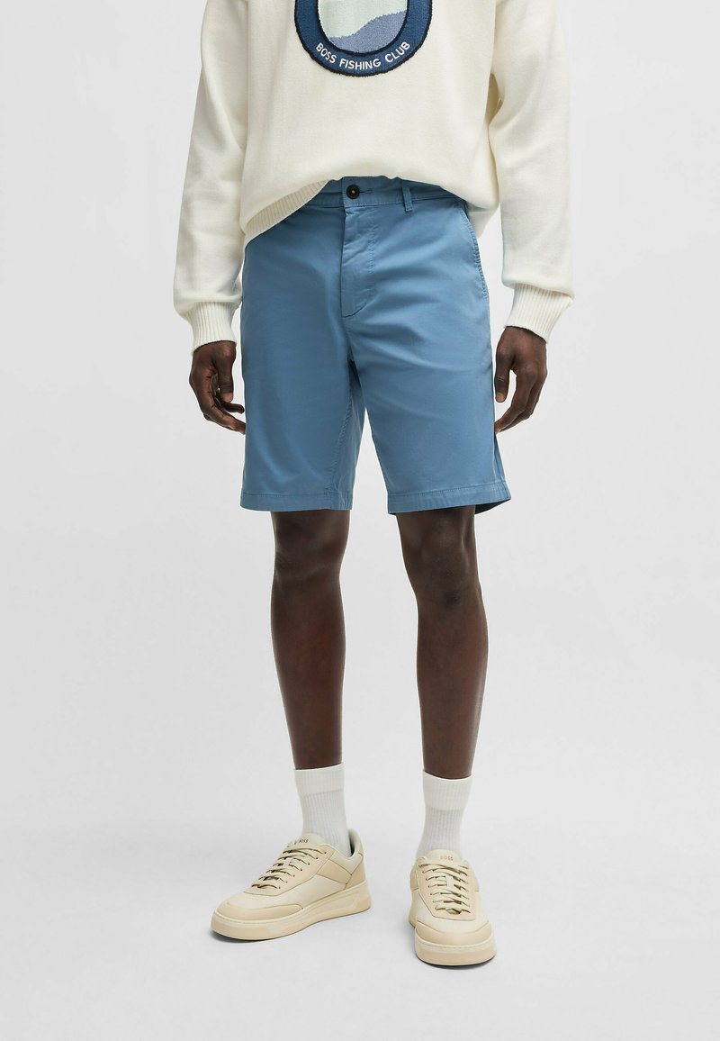 Light blue shorts made of woven fabric, featuring a flat front design, pockets, and a button closure. Worn with a cream sweater and beige sneakers.