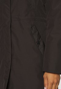 Chaqueta aislante marrón con tejido exterior suave, bolsillo lateral con detalle de botón y costuras reforzadas para mayor durabilidad.