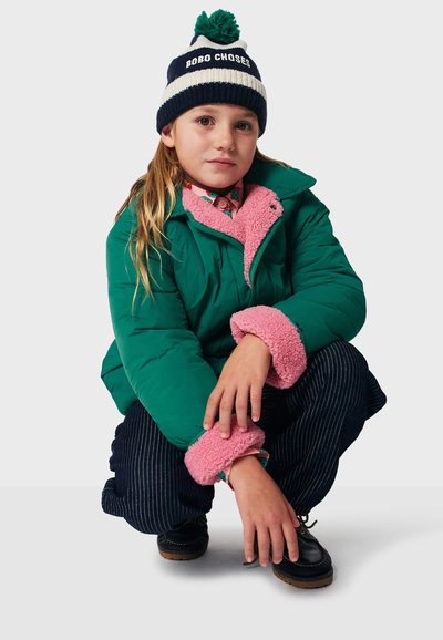 Grön puffjacka med rosa fleecekrage och ärmslut, mönstrad skjorta under, mörka randiga byxor, svarta skor och en marinblå hatt med pom-pom.