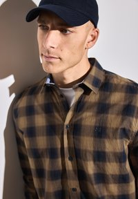 Hnědá a námořnická modrá kostkovaná košile na knoflíky s texturovaným povrchem, doplněná tmavě modrou kšiltovkou, na světlém pozadí.