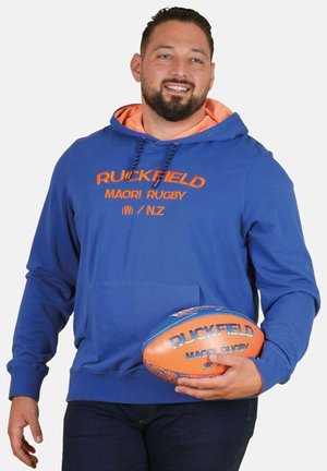 Homme souriant en sweat à capuche bleu tenant un ballon de rugby orange et bleu avec le texte « Ruckfield Maori Rugby » à la fois sur le sweat à capuche et le ballon.