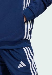 Mornarsko plavi sportski trenirka s bijelim vertikalnim prugama, s prednjim džepom na dukserici i logotipom na hlačama.