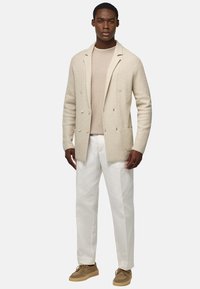 Beige pletený cardigan s dvouřadým zapínáním, v kombinaci s světle hnědým svetrem, bílými kalhotami a béžovými botami, vytvářející vrstvený vzhled.