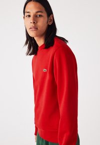 Sweatshirt rouge en tissu doux, avec un col rond, des poignets côtelés et un petit logo vert sur la poitrine.