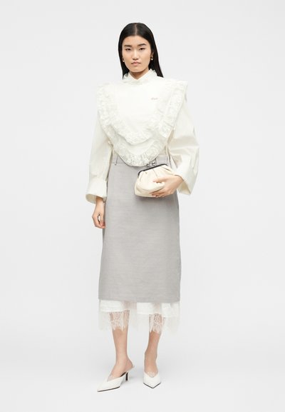 Γυναίκα με λευκό πουκάμισο με δαντελωτές βολάν, γκρι μίντι φούστα, λευκό εσωτερικό φόρεμα με δαντέλα, λευκά τακούνια και κρατώντας μια μπεζ τσάντα φάκελο.