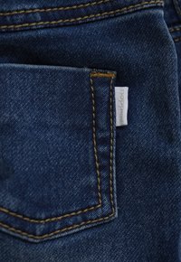 Zbliżenie na tylną kieszeń niebieskich dżinsów z brązowym haftem i małą białą metką marki na prawym brzegu.