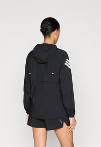 Chaqueta impermeable negra con capucha, bolsillo trasero con cremallera y tres rayas blancas en cada hombro. Textura suave, dobladillo ajustable.